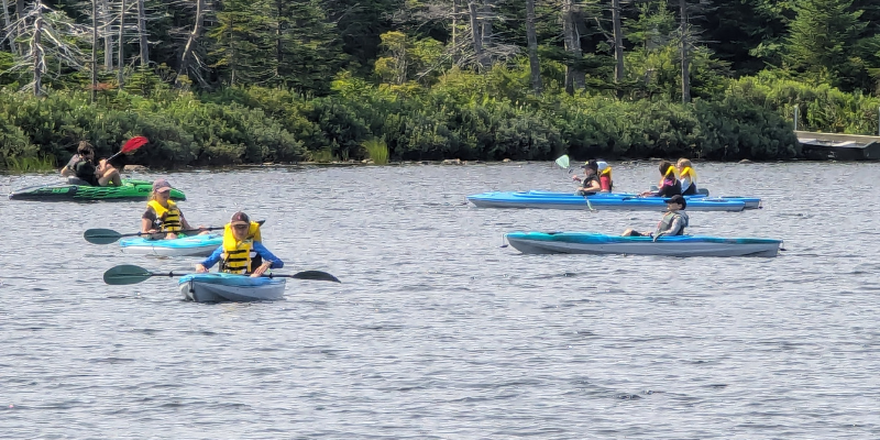 seven children in PFDs paddle kayaks on a pond with an adult supervising